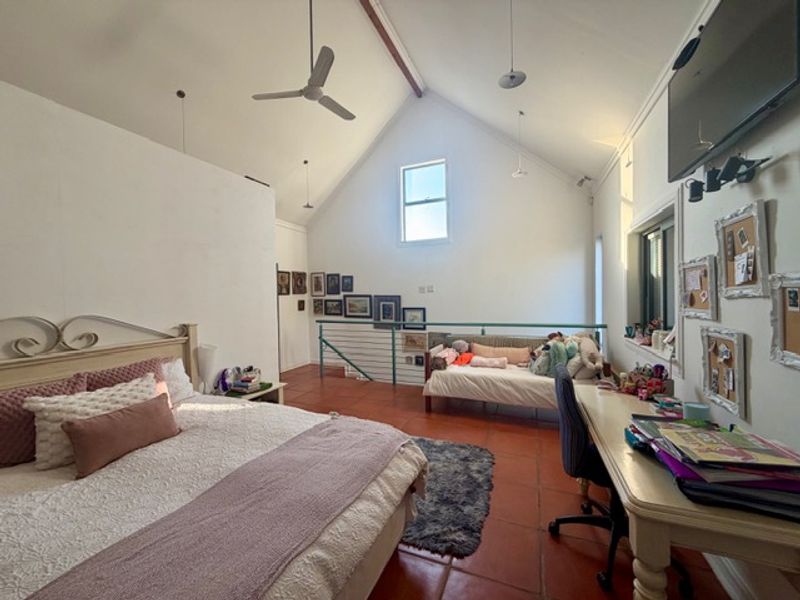 Spacious bedroom featuring a high vaulted ceiling, ceiling fan, terracotta flooring, and a mezzanine level with a metal balustrade.