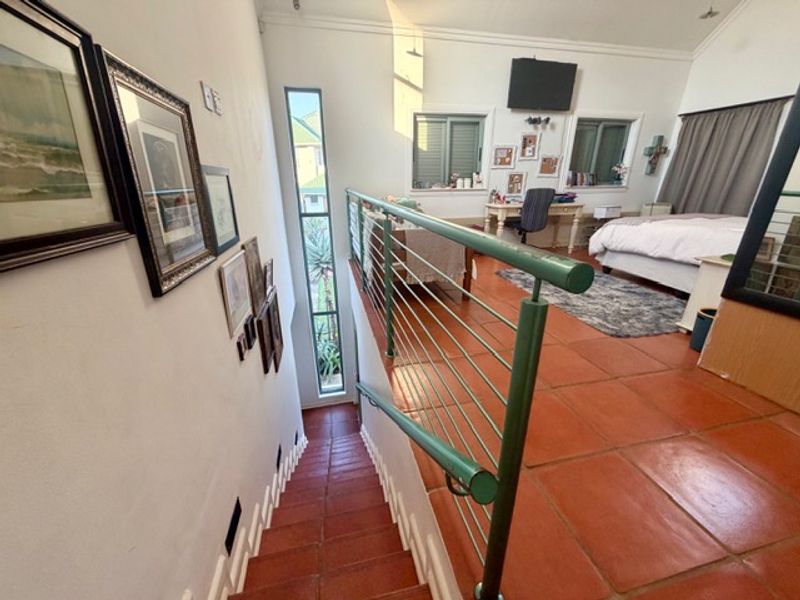 A mezzanine hallway featuring terracotta floor tiles and a metal balustrade, overlooking an open-plan living area with vaulted ceilings.