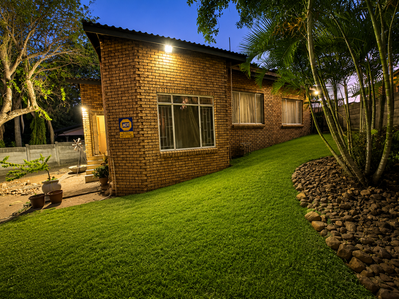 A well-maintained rear exterior view of a face brick house featuring a landscaped lawn, stone retaining wall, and integrated security features at dusk.