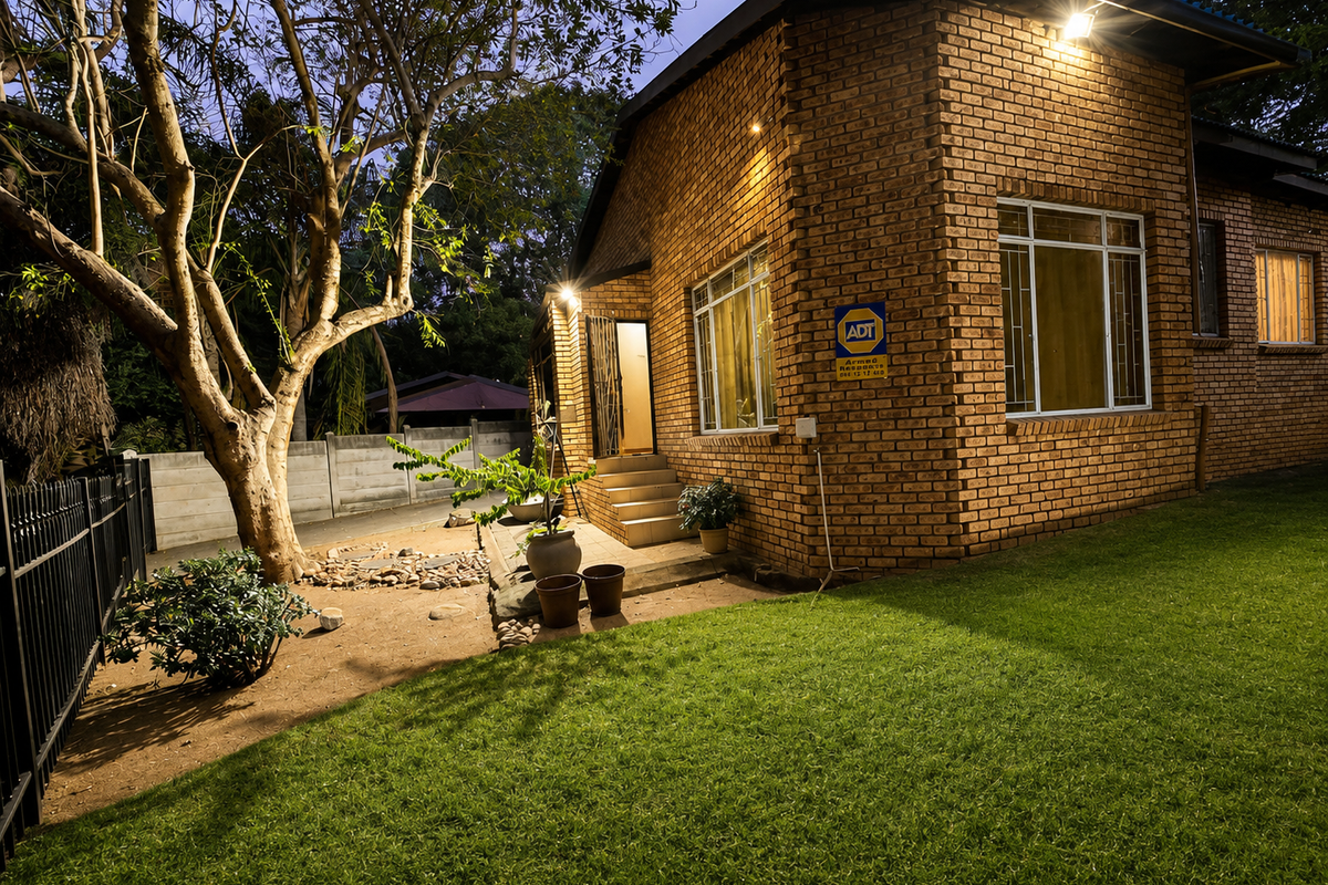 A well-maintained face-brick exterior rear elevation featuring security gates, bars, and outdoor lighting, set against a landscaped garden with a perimeter fence.
