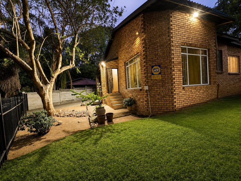 A well-maintained face-brick exterior rear elevation featuring security gates, bars, and outdoor lighting, set against a landscaped garden with a perimeter fence.