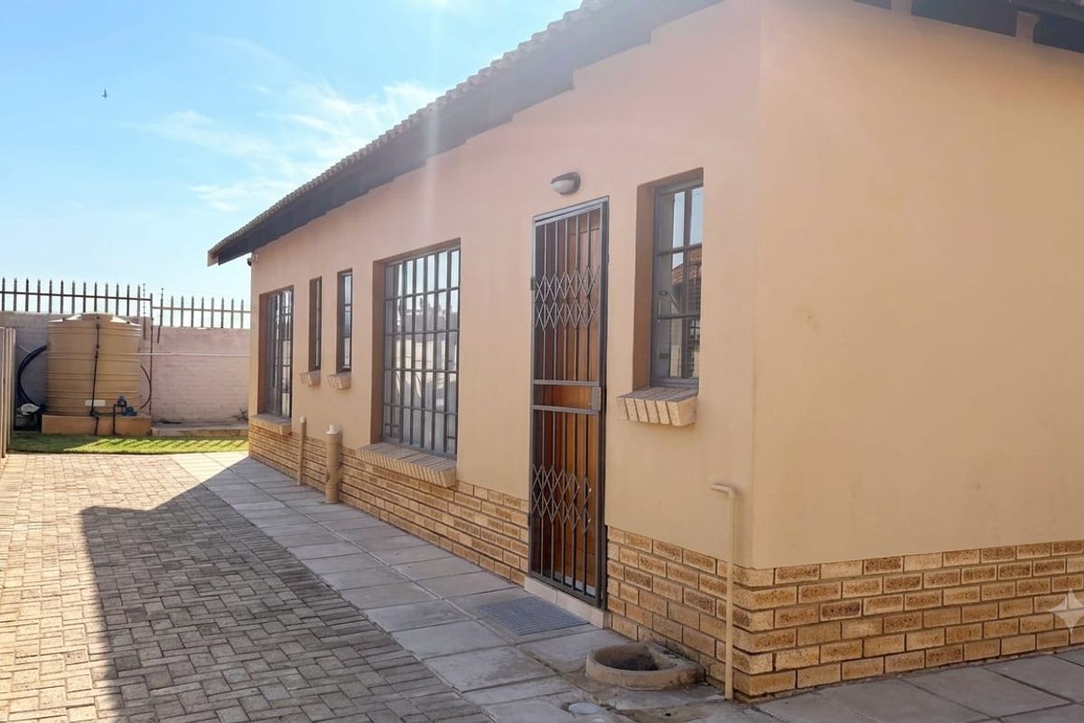 Rear exterior view of the townhouse featuring a paved walkway, security gates, and a water tank situated within a walled perimeter.