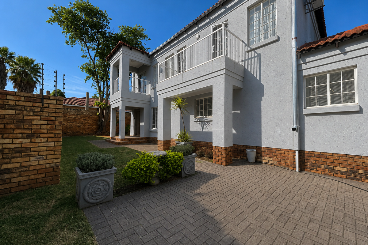 Rear view of a two-story house featuring a paved patio area, a balcony, and a well-maintained garden with brick boundary walls.