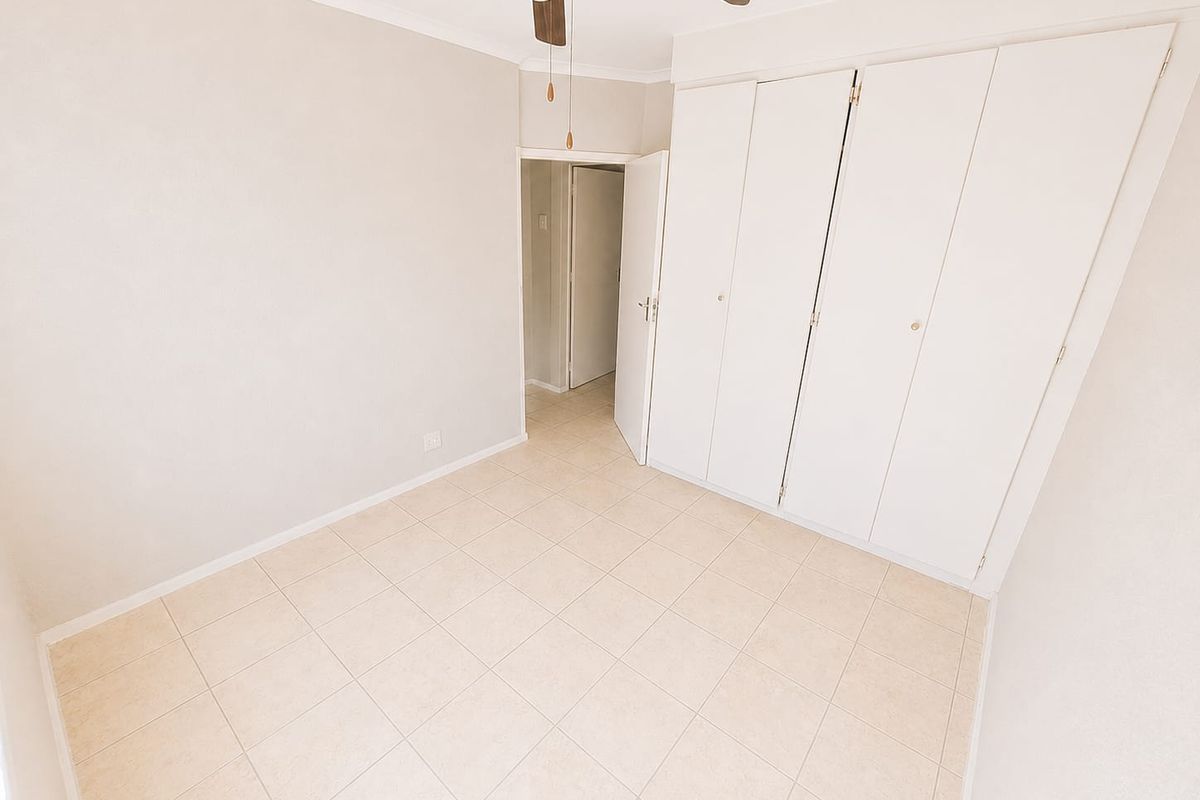 An empty bedroom featuring neutral tiled flooring, built-in cupboards, and a ceiling fan, offering a clean and versatile space.