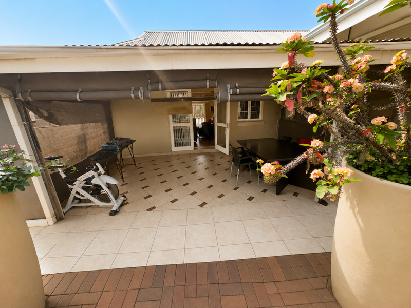 A covered patio area featuring tiled flooring, retractable outdoor blinds, and direct access to the interior through a security gate and sliding door.