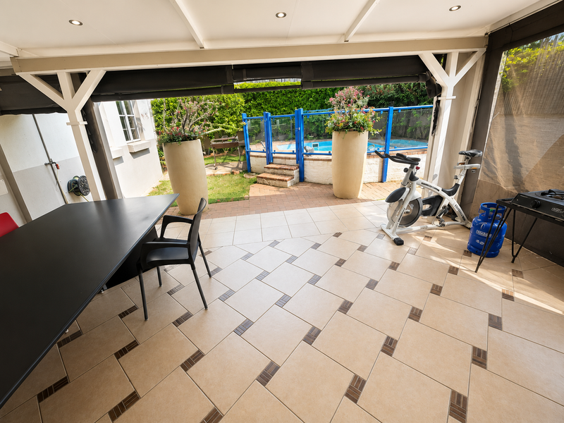 A covered outdoor patio area featuring tiled flooring and recessed lighting, overlooking a private swimming pool with secure fencing and landscaped garden surrounds.