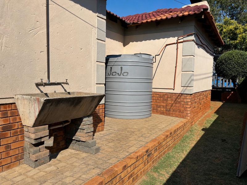 Rear exterior area featuring a water storage tank, a concrete utility sink on a brick base, and paved ground surface.