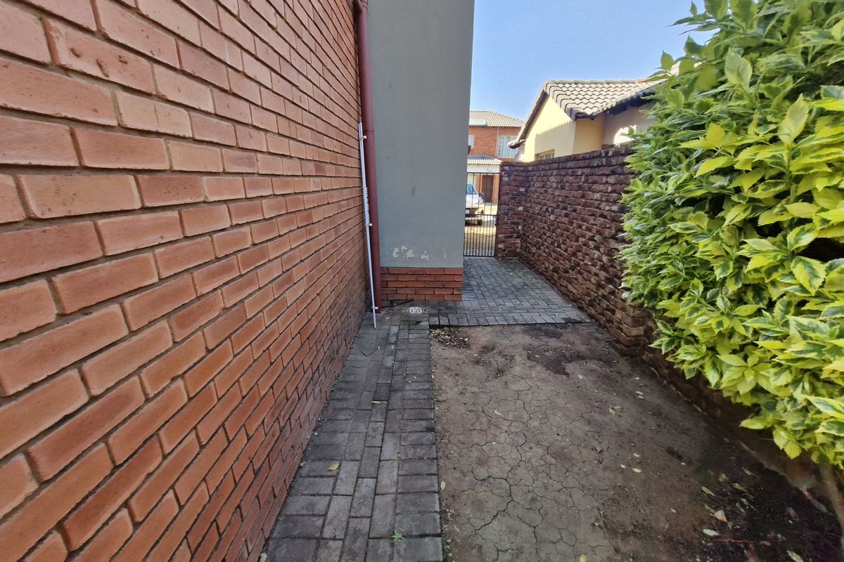 A narrow side passage of the townhouse featuring a brick boundary wall, paved walkway, and a small area of exposed soil.