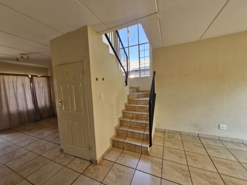 An open-plan living area featuring tiled flooring, a central staircase with a metal balustrade, and a large window providing natural light.