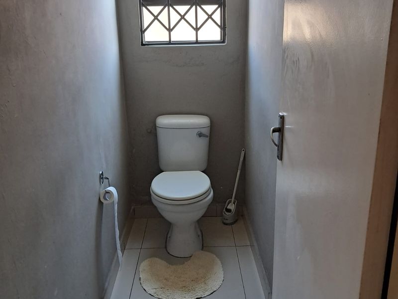 A compact guest toilet featuring tiled flooring and a security-barred window, currently requiring wall finishing to complete the space.
