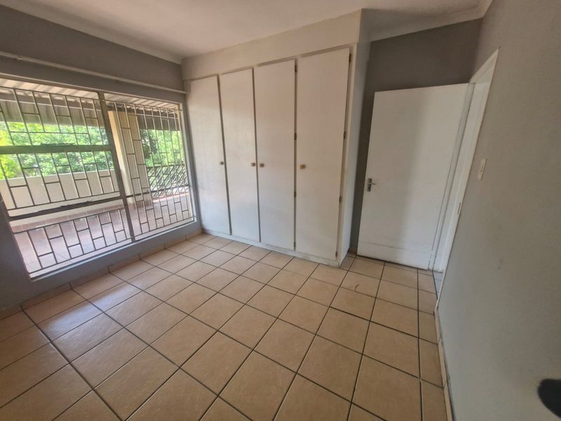 An empty bedroom featuring tiled flooring, built-in cupboards, and a large window with security bars overlooking an outdoor area.