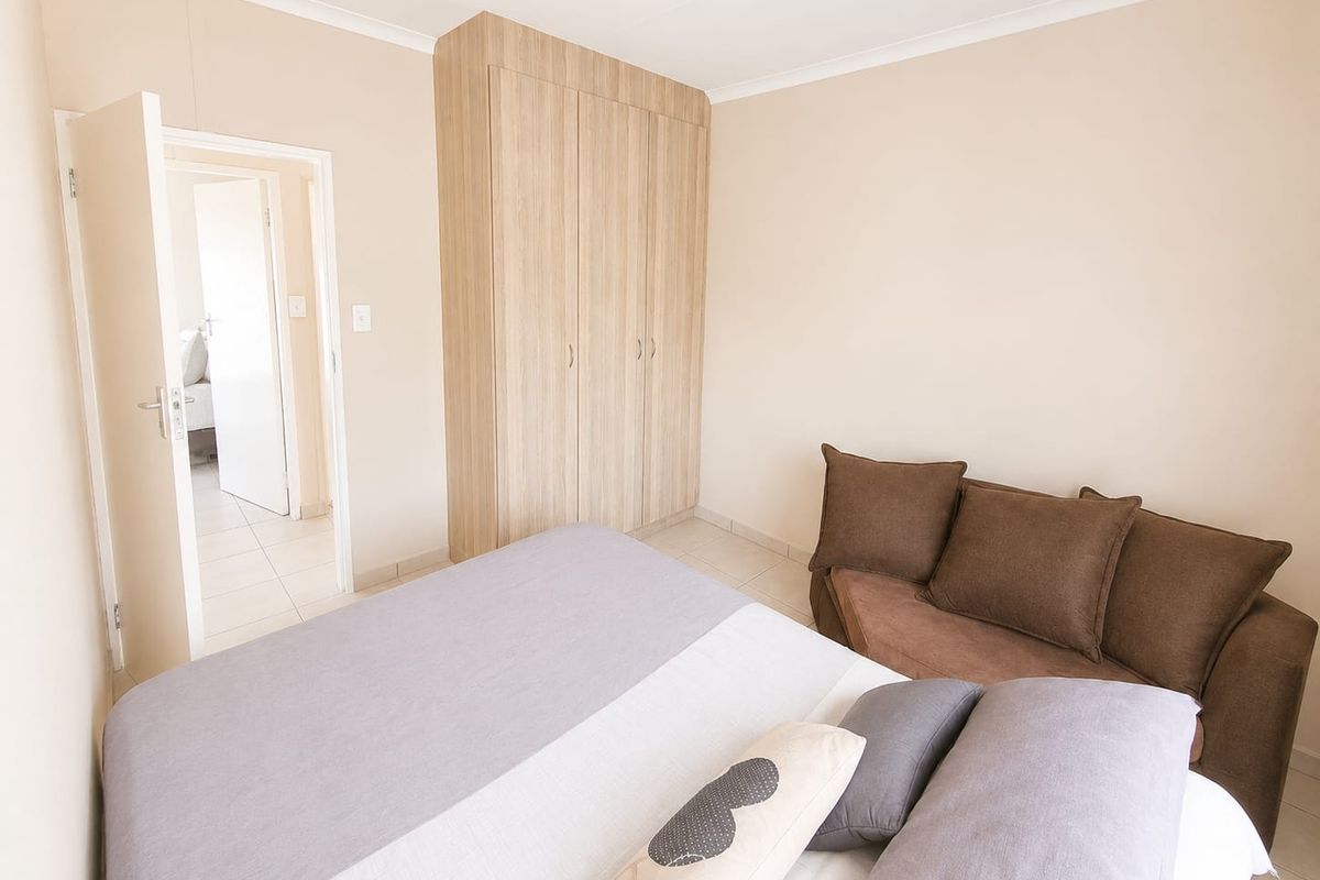 A well-maintained bedroom featuring built-in wooden cupboards, neutral wall finishes, and tiled flooring, with an adjoining doorway leading to another room.