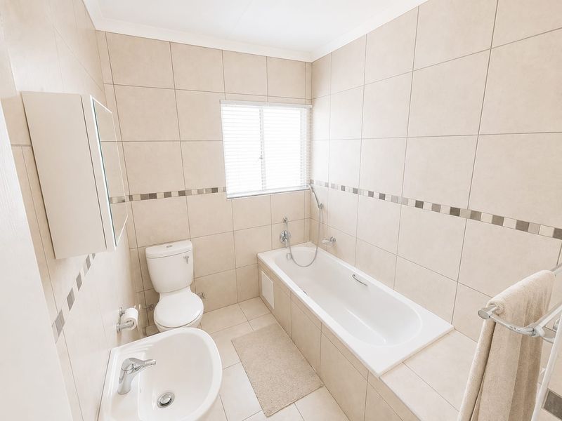A clean, neutral-toned bathroom featuring a built-in bathtub, pedestal basin, toilet, and wall-mounted medicine cabinet with full-height wall tiling.