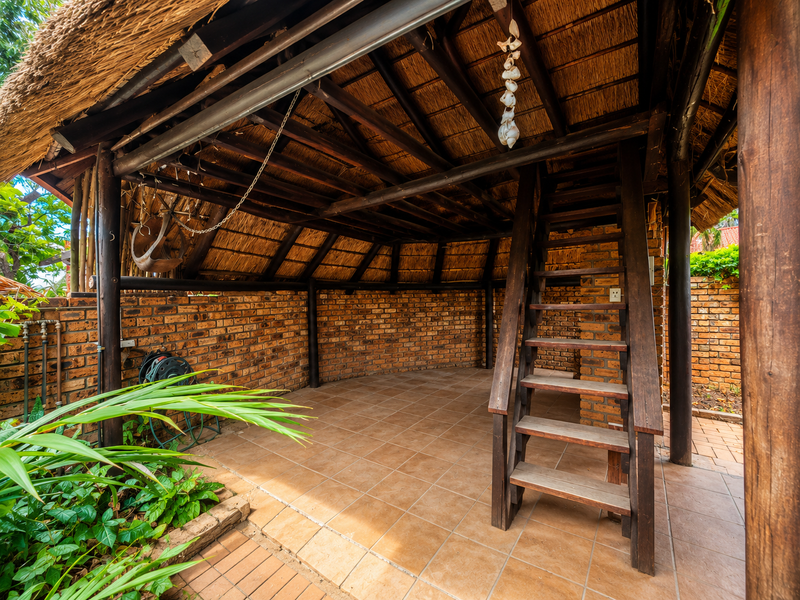 Covered outdoor patio area featuring a rustic thatch roof, exposed timber beams, brick walls, and a wooden staircase leading to an upper level.