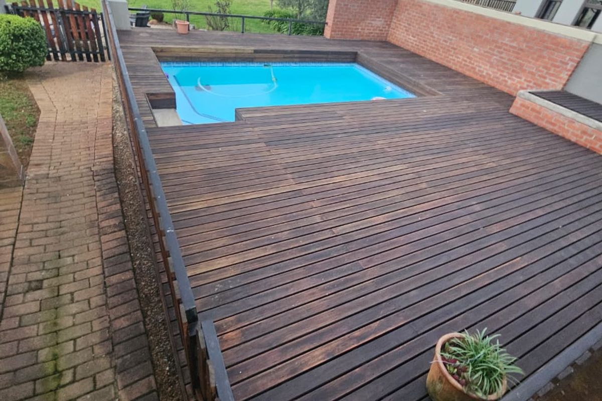 Outdoor pool area featuring a timber deck surround, brick retaining wall, and adjacent brick pathway, overlooking a garden space.