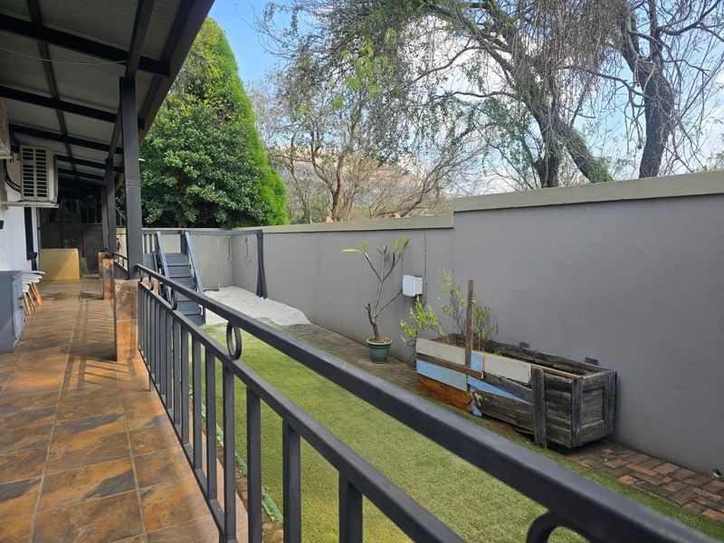 A covered patio area featuring tiled flooring and metal railings, overlooking a small garden space with a boundary wall.