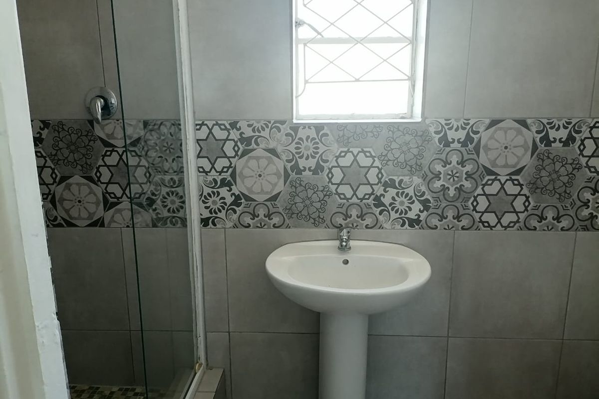 A functional bathroom featuring a pedestal basin, a glass-enclosed shower, and decorative patterned wall tiling with a window for natural light.