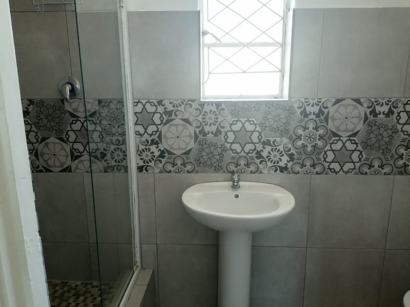 A functional bathroom featuring a pedestal basin, a glass-enclosed shower, and decorative patterned wall tiling with a window for natural light.