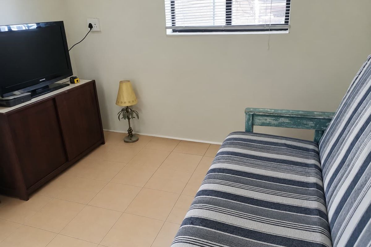 A simple living room area featuring neutral tiled flooring, a window with venetian blinds, and standard electrical fixtures.