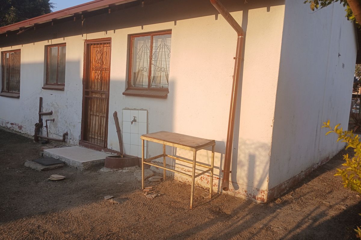 Rear exterior view of the house showing exposed plumbing, security gate, and signs of wear on the exterior wall finish.