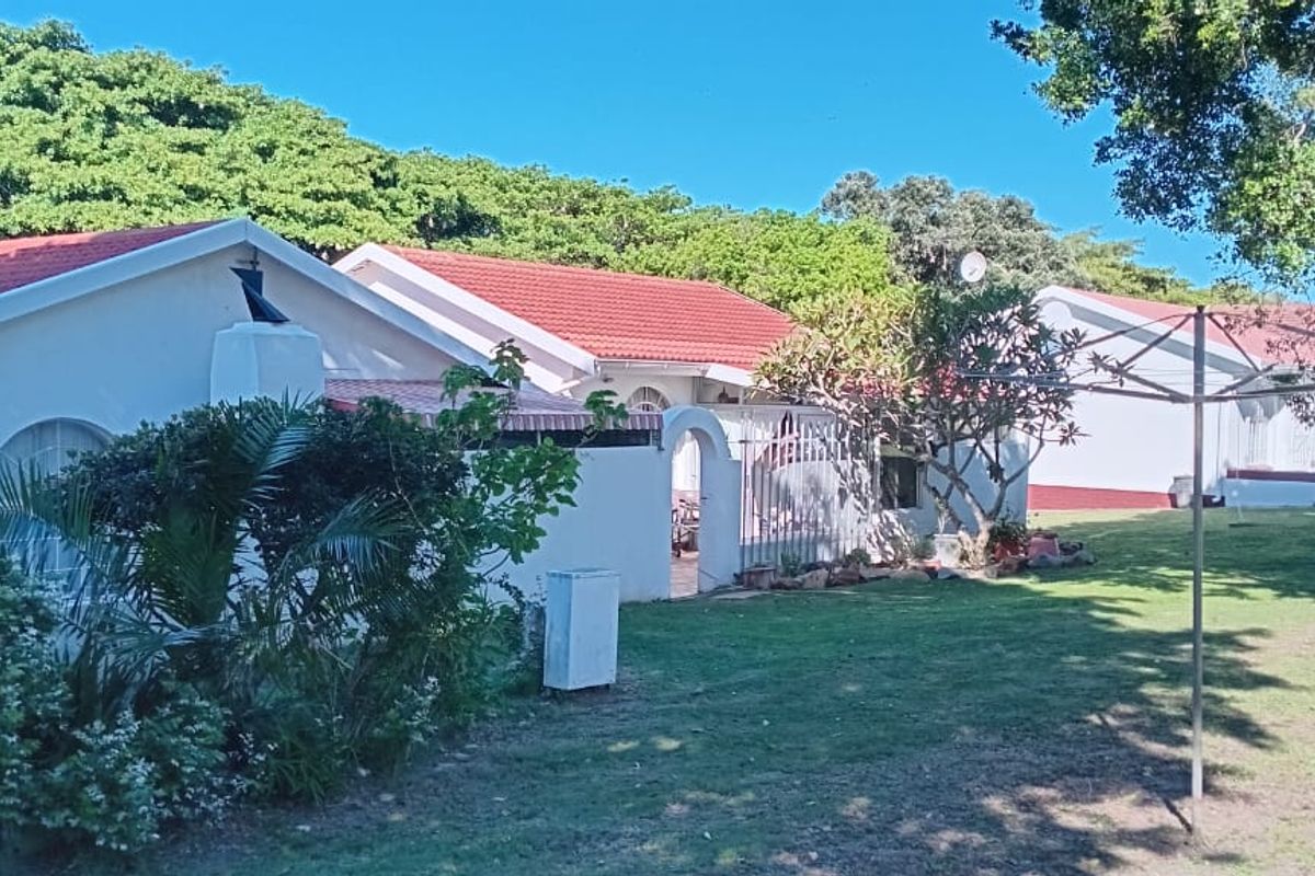 Rear view of the townhouse complex featuring plastered walls, tiled roofing, a shared lawn area, and a built-in outdoor braai facility.
