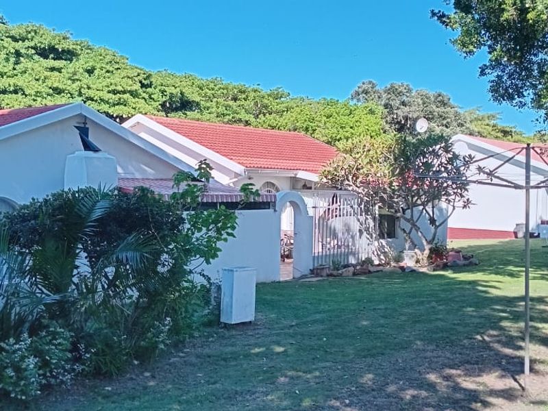 Rear view of the townhouse complex featuring plastered walls, tiled roofing, a shared lawn area, and a built-in outdoor braai facility.