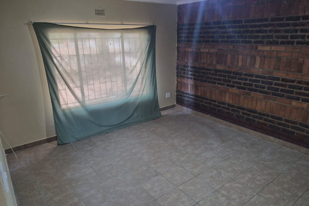 An empty living room featuring a prominent exposed brick wall, tiled flooring, and a window fitted with security bars.