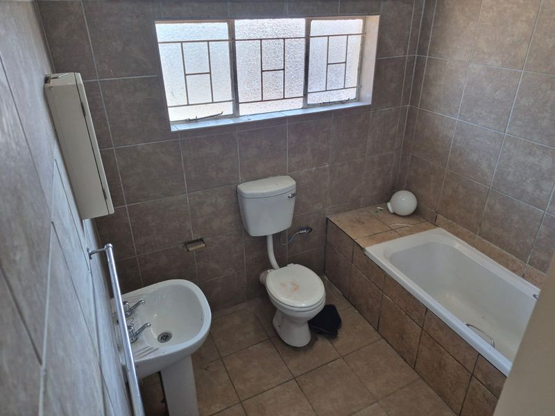 A dated bathroom featuring a pedestal basin, toilet, and built-in bathtub with full-height wall tiling and security bars on the window.