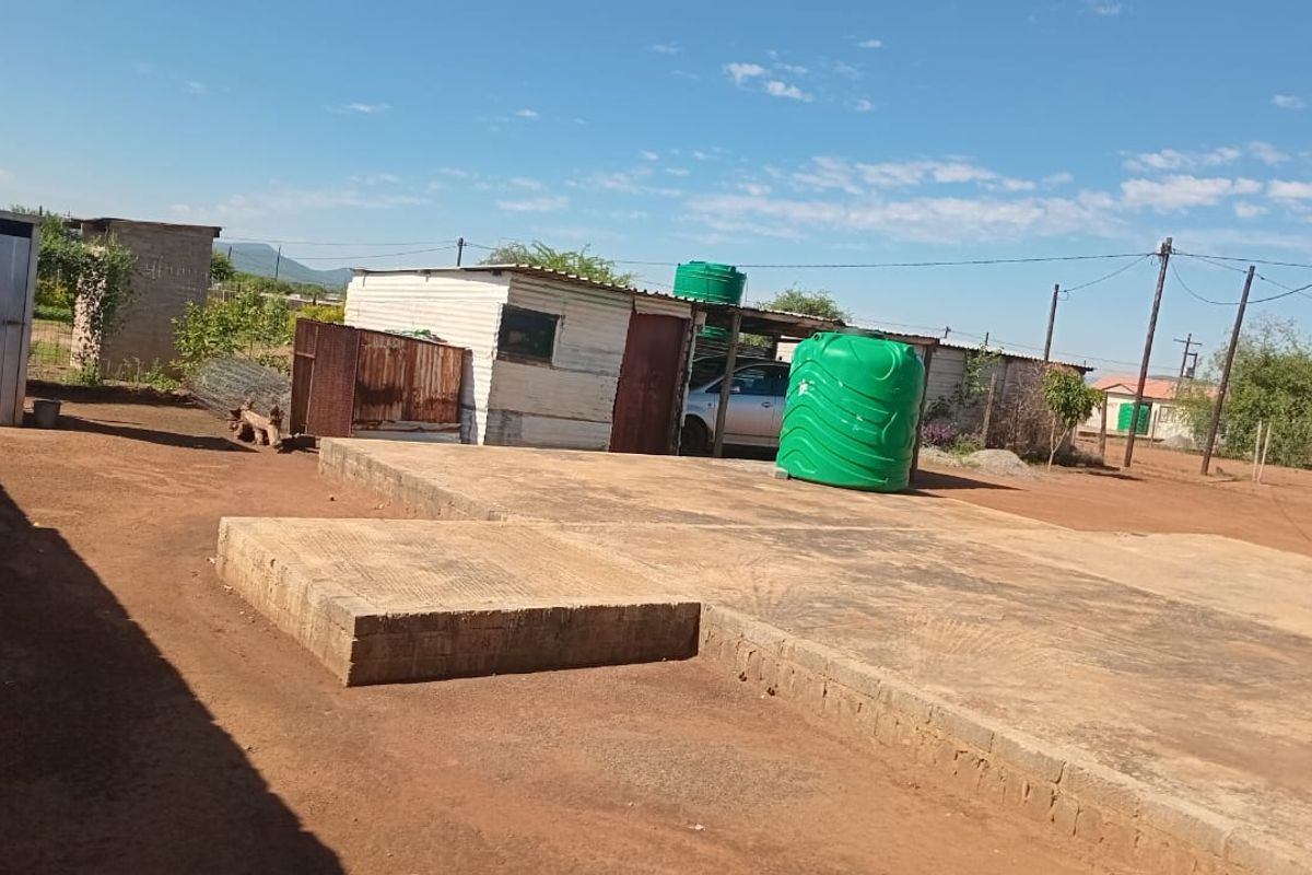 Rear yard area featuring a concrete slab foundation, corrugated metal outbuilding, and a plastic water storage tank on a dirt plot.