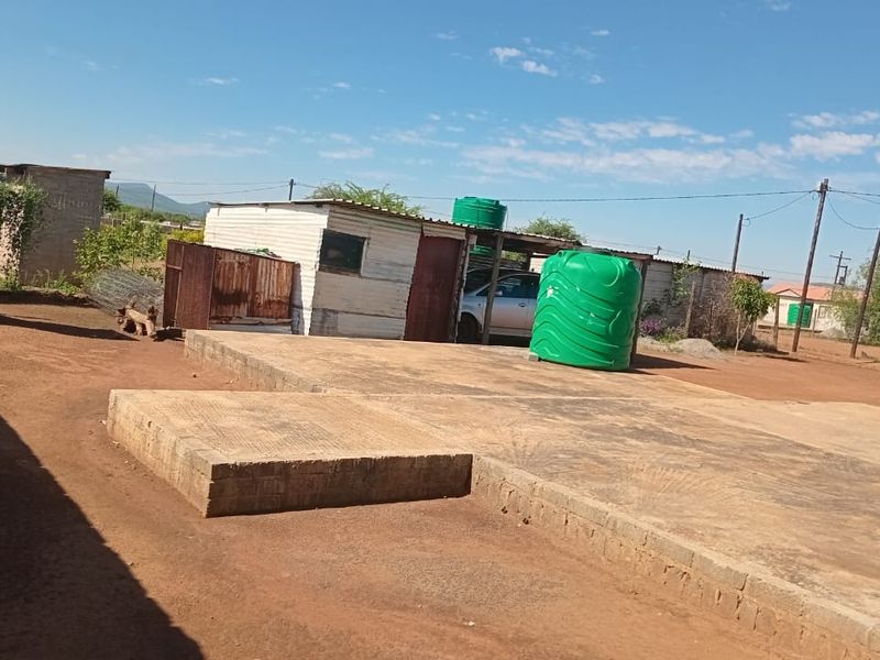 Rear yard area featuring a concrete slab foundation, corrugated metal outbuilding, and a plastic water storage tank on a dirt plot.