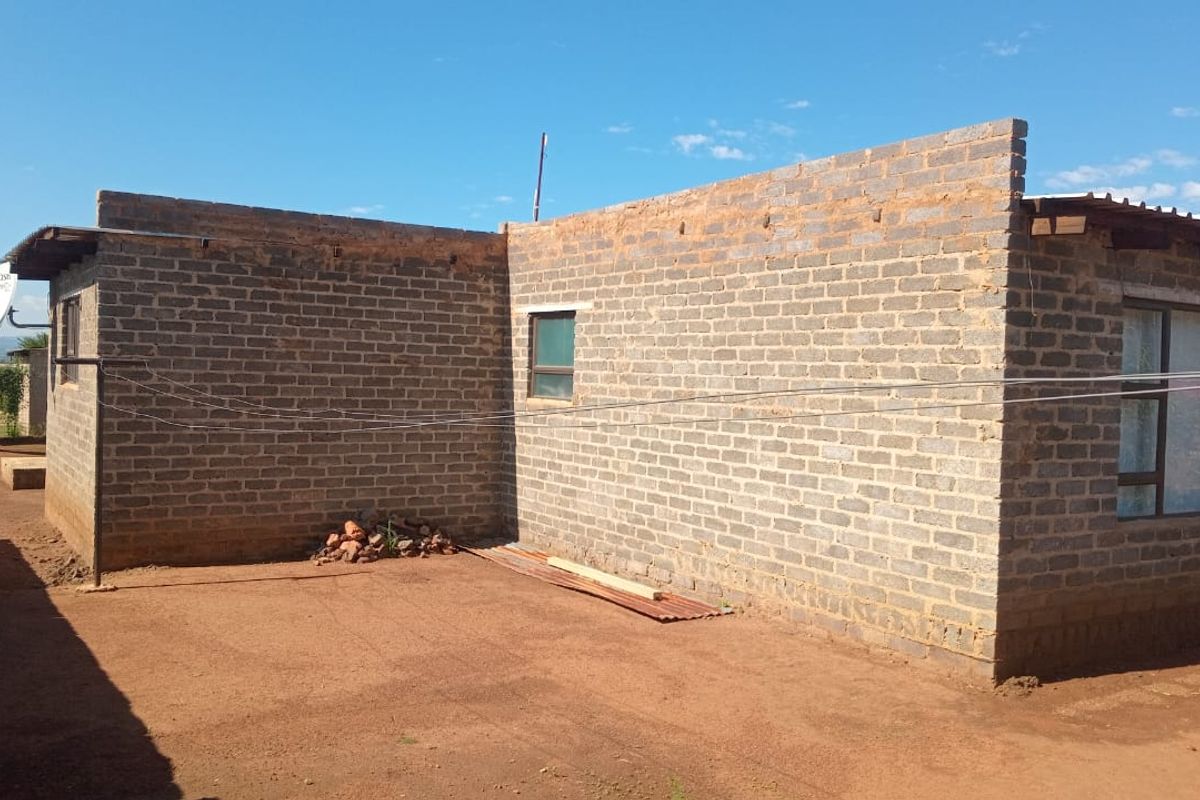 Rear view of a house featuring exposed brick walls, aluminium window frames, and a corrugated metal roof, set on a dirt yard.