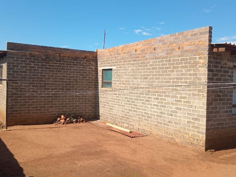 Rear view of a house featuring exposed brick walls, aluminium window frames, and a corrugated metal roof, set on a dirt yard.