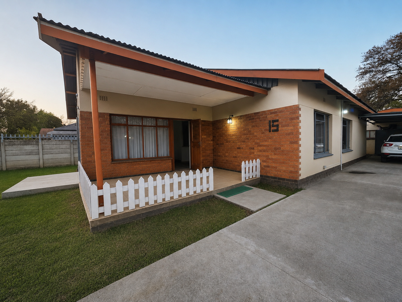 A well-maintained single-story house featuring a brick facade, a covered front patio with a picket fence, and a spacious concrete driveway.