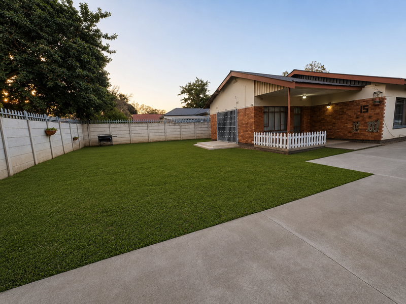 A well-maintained rear exterior featuring a spacious artificial lawn, concrete driveway, and a covered patio area with brick facade detailing.