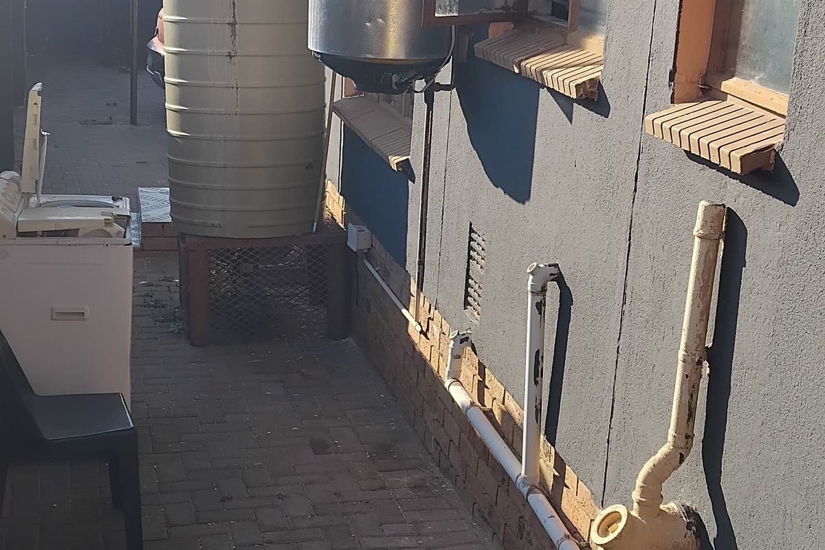 Rear exterior view showing paved walkway, external plumbing, water storage tank, and wall-mounted electric geyser unit.