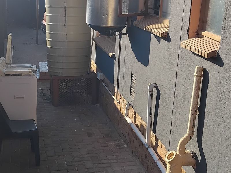 Rear exterior view showing paved walkway, external plumbing, water storage tank, and wall-mounted electric geyser unit.