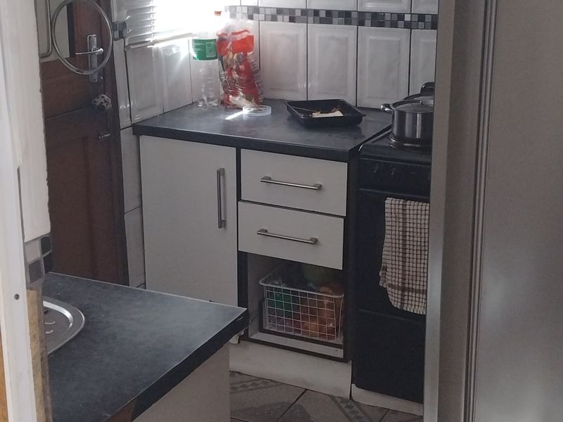 A compact kitchen featuring white cabinetry, dark laminate countertops, tiled backsplash, and ceramic flooring, equipped with a freestanding stove and extractor fan.
