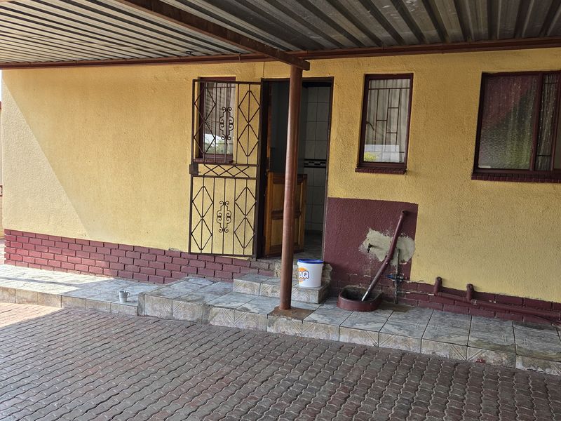 Front exterior view featuring a covered patio area with paved driveway, security gate, and visible wall maintenance requirements.