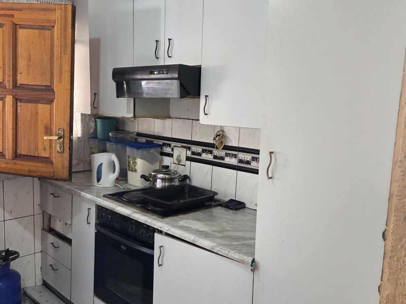 A compact kitchen featuring white built-in cabinetry, a built-in oven with hob, extractor fan, and tiled finishes, currently showing signs of wear and maintenance needs.