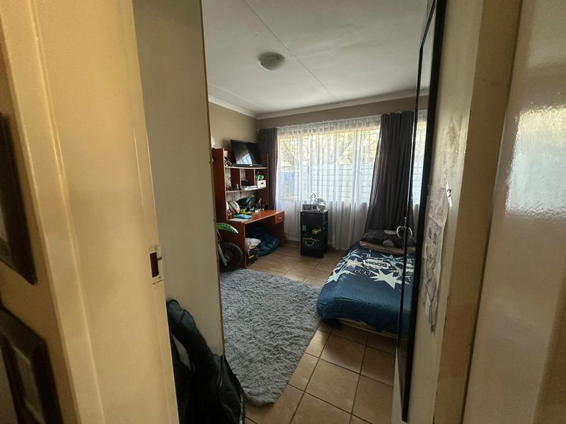 A compact bedroom featuring tiled flooring and a large window with curtain rails, offering a functional space for a single bed and study area.