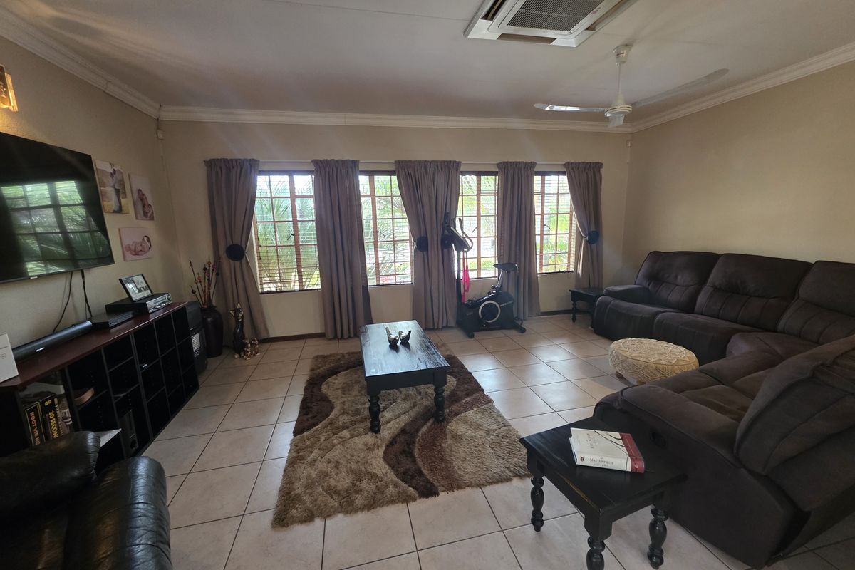 Spacious living room featuring tiled flooring, coved cornicing, a ceiling fan, and ducted air conditioning, with large windows fitted with security bars.