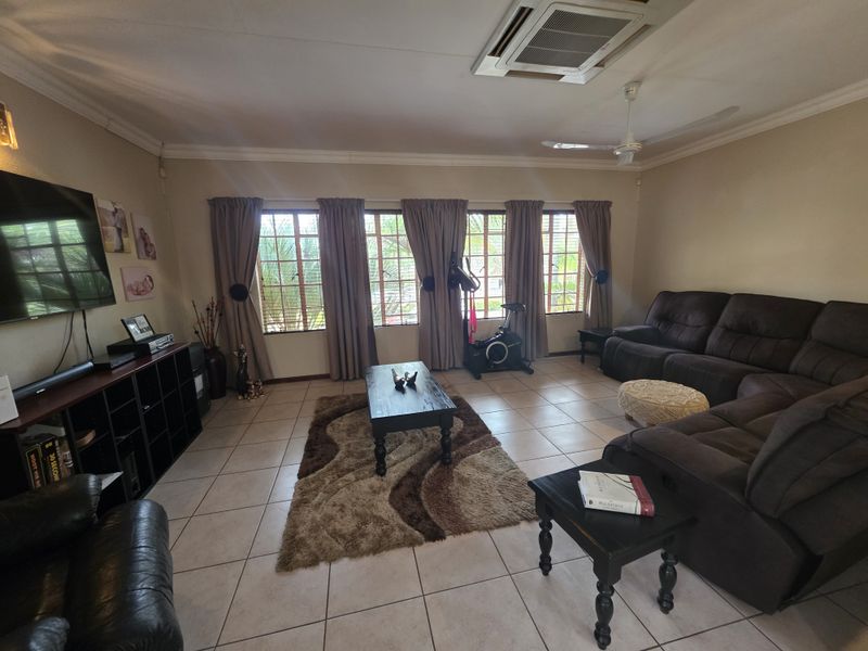 Spacious living room featuring tiled flooring, coved cornicing, a ceiling fan, and ducted air conditioning, with large windows fitted with security bars.