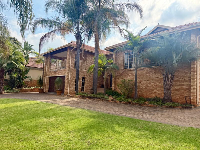 A well-maintained double-story brick house featuring a paved driveway, established palm trees, and a balcony overlooking the front garden.