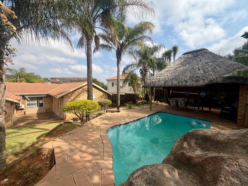Outdoor pool area featuring a swimming pool, paved surround, and a thatched lapa, set within a landscaped garden with mature palm trees.