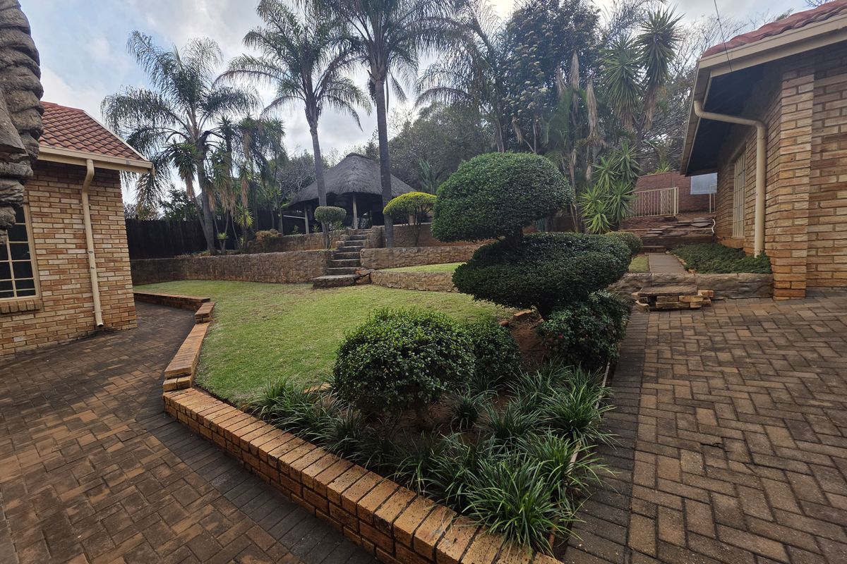 A well-maintained rear garden featuring brick paving, tiered retaining walls, landscaped shrubbery, and a thatched gazebo area.
