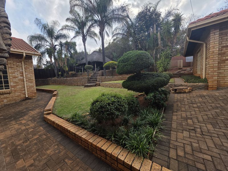 A well-maintained rear garden featuring brick paving, tiered retaining walls, landscaped shrubbery, and a thatched gazebo area.