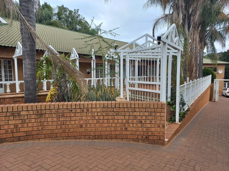 Front exterior view of a brick house featuring a paved driveway, metal pergola entrance, and established garden beds with a brick boundary wall.