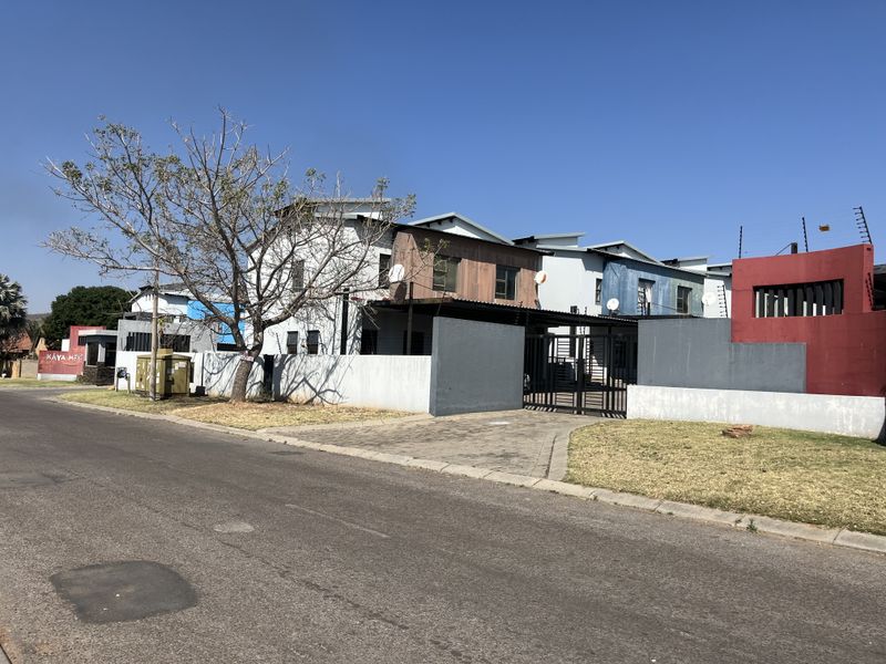 Front exterior view of a townhouse complex featuring a paved driveway, perimeter wall with electric fencing, and multi-colored facade finishes.