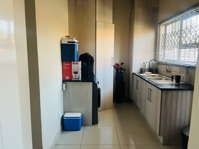 A functional scullery featuring light wood-finish cabinetry, a double basin sink, tiled floors, and a window with security bars.
