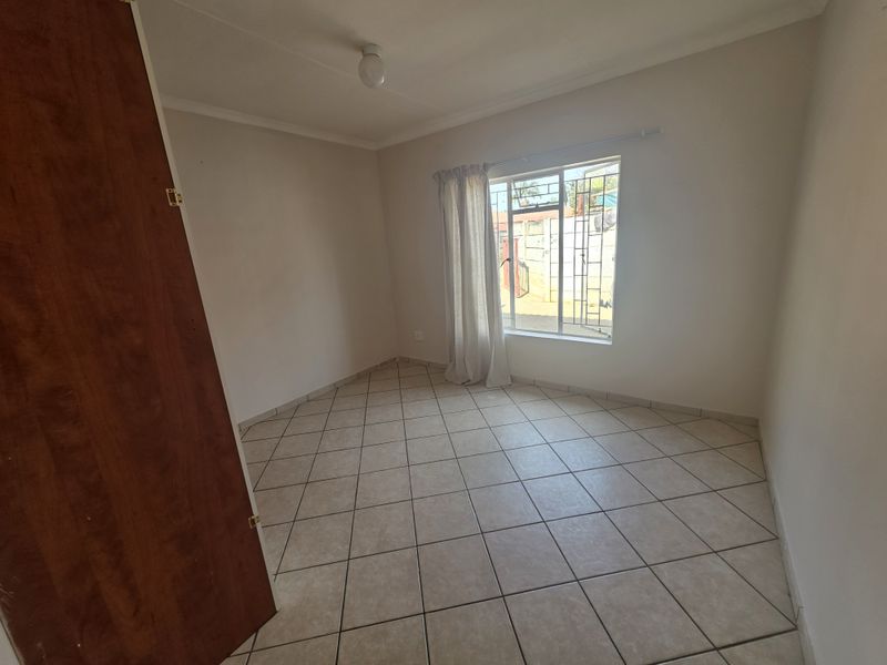 A bright, empty bedroom featuring neutral tiled flooring, a built-in cupboard, and a window with security bars.